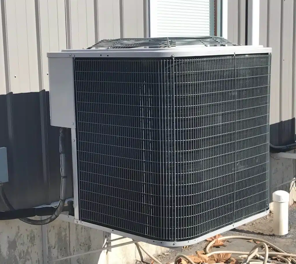 A gray outdoor air conditioner with leaves underneath it